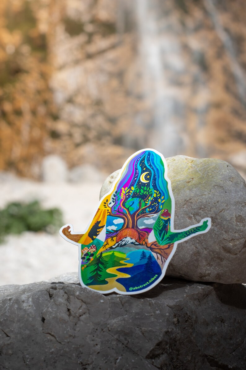 Puede incluir: Una pegatina colorida que representa una figura humana estilizada con un &aacute;rbol que crece de su pecho. La figura est&aacute; rodeada de escenas de la naturaleza, incluyendo monta&ntilde;as, un r&iacute;o y un bosque. La pegatina est&aacute; en una roca gris con una cascada borrosa en el fondo. El texto "@abstract.hikes" est&aacute; en la parte inferior de la pegatina.