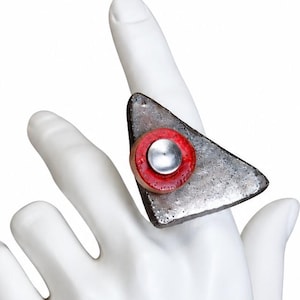 May include: A ring featuring a large, triangular, textured silver-toned metal plate. Centered on the plate is a circular red element with a smaller, smooth, metallic silver button-like detail at its core, creating a layered, geometric design.