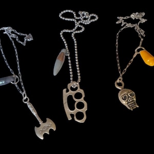 May include: Three silver pendant necklaces on a black backdrop. One has a small axe, another a set of brass knuckles, and the third a skull. Each necklace has a different chain style and a unique charm.