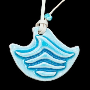 May include: A light blue ceramic pendant with a wave design, strung on a white leather cord. The pendant has a crackle glaze finish and a small turquoise bead. The background is black.
