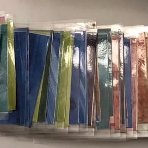 Può includere: Una collezione di fogli di carta rettangolari colorati in vari colori, tra cui viola, rosso, oro, verde e blu, sono disposti in fila. Le carte sono in buste di plastica trasparente e i colori variano da tinta unita a metallizzato.
