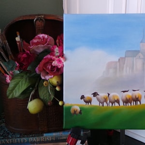 May include: A painting of a church with a steeple and a row of sheep in front of it. The painting is on an easel and there are paintbrushes and flowers in a wooden box to the left.