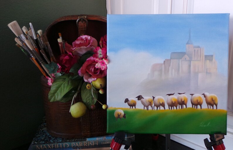May include: A painting of a church with a steeple and a row of sheep in front of it. The painting is on an easel and there are paintbrushes and flowers in a wooden box to the left.