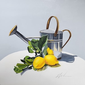 May include: A silver watering can with a brass spout and handle sits on a white surface with three yellow lemons and green leaves.