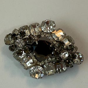 Puede incluir: Broche vintage con una piedra central oscura de forma ovalada, rodeada de pedrería transparente y gris ahumado. El broche, con forma de diamante, está engastado sobre un reverso de metal plateado. Las piedras son de varias formas y tamaños.