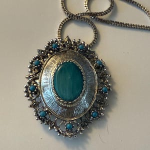 May include: Silver-toned pendant necklace with a large oval turquoise-colored stone at the center. The pendant is surrounded by a decorative silver frame with small turquoise-colored accents and a matching chain.