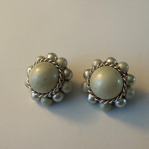 May include: A pair of vintage clip-on earrings. Each earring features a large, pale green central bead surrounded by a ring of smaller, matching beads. A silver-tone chain encircles the central bead. The earrings are set against a plain, off-white background.