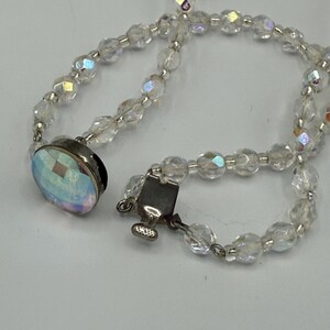 May include: A clear, iridescent beaded necklace with a silver clasp and a large, oval-shaped pendant. The necklace is composed of small, faceted beads that catch the light, creating a shimmering effect. The pendant has a similar iridescent finish.