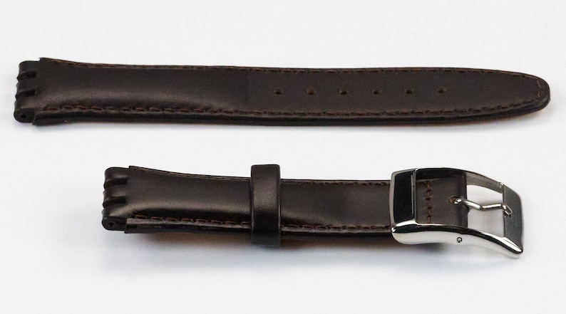 K&ouml;nnte beinhalten: Ein braunes Lederarmband mit einer silbernen Schlie&szlig;e. Das Armband hat einen gen&auml;hten Rand und ist f&uuml;r eine Uhr mit einer Bandansto&szlig;breite von 18 mm ausgelegt.