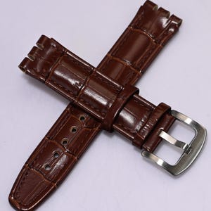 May include: Brown leather watch band with a crocodile skin texture. The band features a silver-colored buckle and multiple adjustment holes. The watch band is designed to be durable and stylish.