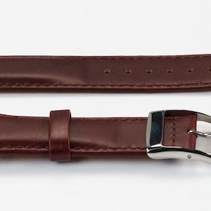 K&ouml;nnte beinhalten: Ein braunes Lederarmband mit einer silbernen Schlie&szlig;e. Das Armband hat ein strukturiertes Design und ist mit wei&szlig;em Garn vern&auml;ht.