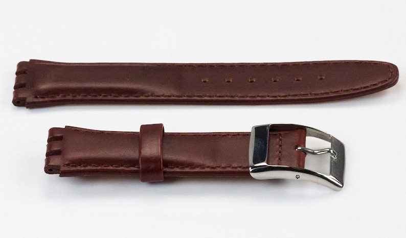 K&ouml;nnte beinhalten: Ein braunes Lederarmband mit einer silbernen Schlie&szlig;e. Das Armband hat ein strukturiertes Design und ist mit wei&szlig;em Garn vern&auml;ht.