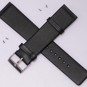 May include: Black leather watch strap with a silver buckle and several small screws. The strap has a textured surface and is designed to attach to a watch face. The strap is shown against a white background.
