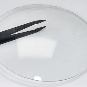 May include: A transparent, circular lens with a black tweezer resting on top. The lens is clear, allowing light to pass through. The tweezer is made of a dark material and is positioned to hold the lens. The image is set against a white background.