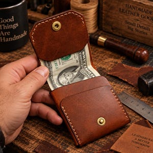 Puede incluir: Una cartera de cuero marrón con solapa plegable y cierre de botón a presión de latón. La cartera presenta costuras visibles de color claro a lo largo de los bordes y contiene un billete de un dólar estadounidense dentro del compartimento.