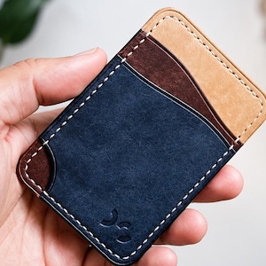 May include: A rectangular leather card holder featuring a tri-colour design with a navy blue central pocket, a dark brown side pocket, and a light tan top pocket. The wallet is constructed with visible white stitching and a small embossed logo.