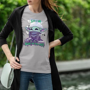 May include: A light gray t-shirt featuring a cartoon character with large ears, wearing a purple scarf and a helmet. The shirt has the text "Cute I am" and "Adore me you must". The person is wearing blue jeans and a black cardigan.