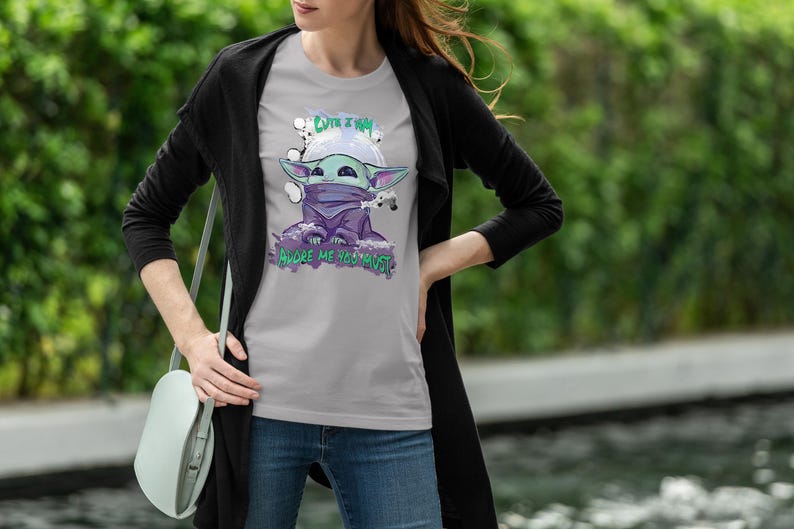 May include: A light gray t-shirt featuring a cartoon character with large ears, wearing a purple scarf and a helmet. The shirt has the text "Cute I am" and "Adore me you must". The person is wearing blue jeans and a black cardigan.