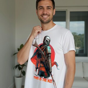 Op de afbeelding: Wit t-shirt met een afbeelding van een personage met helm, geweer en cape tegen een rode en oranje achtergrond. De tekst "THIS IS THE WAY" staat eronder gedrukt. De persoon glimlacht.