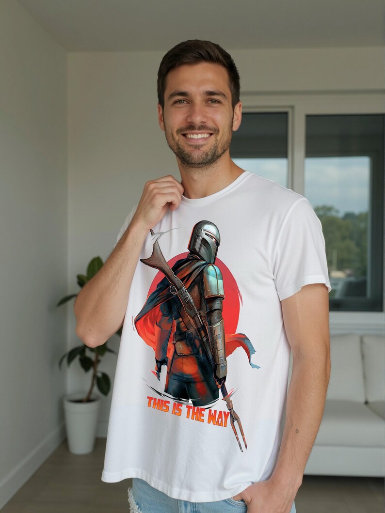 Op de afbeelding: Wit t-shirt met een afbeelding van een personage met helm, geweer en cape tegen een rode en oranje achtergrond. De tekst "THIS IS THE WAY" staat eronder gedrukt. De persoon glimlacht.
