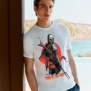 Op de afbeelding: Wit T-shirt met een afbeelding van een Mandalorian-personage tegen een rode zonsopgang. De tekst "THIS IS THE WAY" is in het rood weergegeven. Het T-shirt wordt door een persoon gedragen.