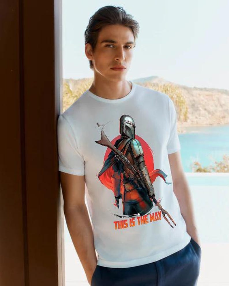 Op de afbeelding: Wit T-shirt met een afbeelding van een Mandalorian-personage tegen een rode zonsopgang. De tekst "THIS IS THE WAY" is in het rood weergegeven. Het T-shirt wordt door een persoon gedragen.