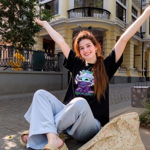 May include: A black t-shirt with a graphic of a cartoon character, light blue pants, and beige platform sandals. A floral tote bag rests on the ground. The person is sitting on a brick surface with arms raised. A building is in the background.