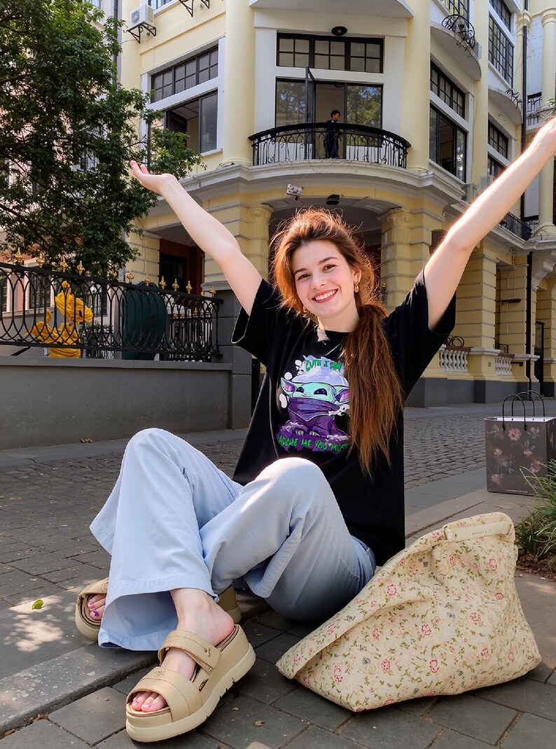 May include: A black t-shirt with a graphic of a cartoon character, light blue pants, and beige platform sandals. A floral tote bag rests on the ground. The person is sitting on a brick surface with arms raised. A building is in the background.