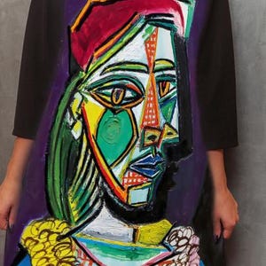 May include: A black dress featuring a large, colorful, cubist-style portrait print on the front. The artwork displays a face in shades of green, yellow, and blue with a red hat, set against a deep purple background with a checkered pattern at the base.
