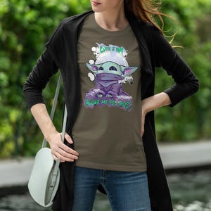 May include: Olive green t-shirt featuring a cartoon character with large ears, a purple cloak, and a face covering. The text "CUTE I AM" is above the character, and "ADORE ME YOU MUST" is below. The shirt is worn by a person.