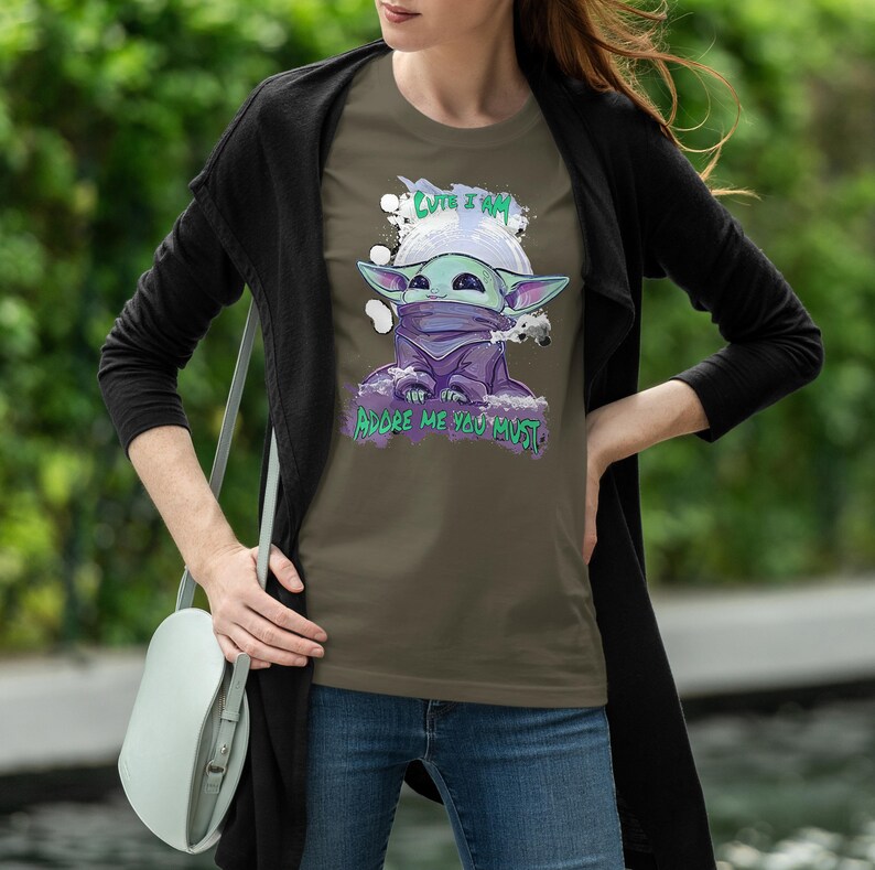 May include: Olive green t-shirt featuring a cartoon character with large ears, a purple cloak, and a face covering. The text "CUTE I AM" is above the character, and "ADORE ME YOU MUST" is below. The shirt is worn by a person.
