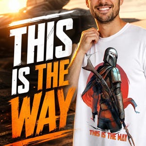 Op de afbeelding: Wit t-shirt met een afbeelding van een personage in harnas met een wapen, met de tekst "THIS IS THE WAY". De achtergrond toont een zonsondergang en een ruimteschip. De persoon glimlacht.