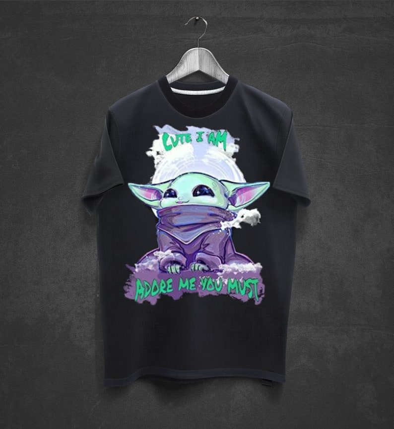 May include: Black t-shirt featuring a cartoon illustration of a green alien-like character with large ears and a purple cloak. The text "CUTE I AM" and "ADORE ME YOU MUST" are above and below the character, respectively.