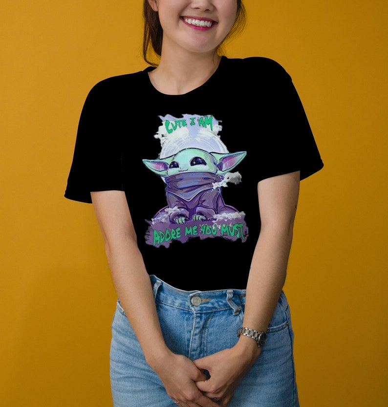 May include: Black t-shirt featuring a cartoon character with large ears, a purple cloak, and the text "CUTE I AM" and "ADORE ME YOU MUST" in green and white. The character is set against a white and purple background.