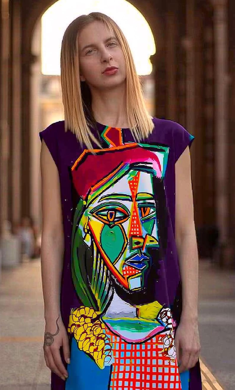 May include: A sleeveless purple tunic dress featuring a large, colorful, abstract cubist-style portrait graphic on the front. The design incorporates vibrant shades of green, yellow, red, and blue in a geometric pattern with bold black outlines.