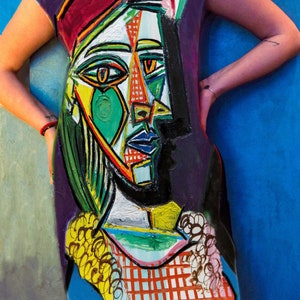 May include: A sleeveless dress featuring a vibrant, abstract cubist-style portrait print. The design incorporates bold blocks of green, red, yellow, and blue, with thick black outlines and a checkered pattern at the bottom hem on a dark purple background.