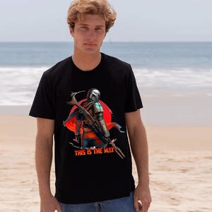 Op de afbeelding: Zwart t-shirt met een afbeelding van een personage met helm en wapen, tegen een rode cirkel. De tekst "THIS IS THE WAY" staat eronder. Het t-shirt wordt door een persoon gedragen.