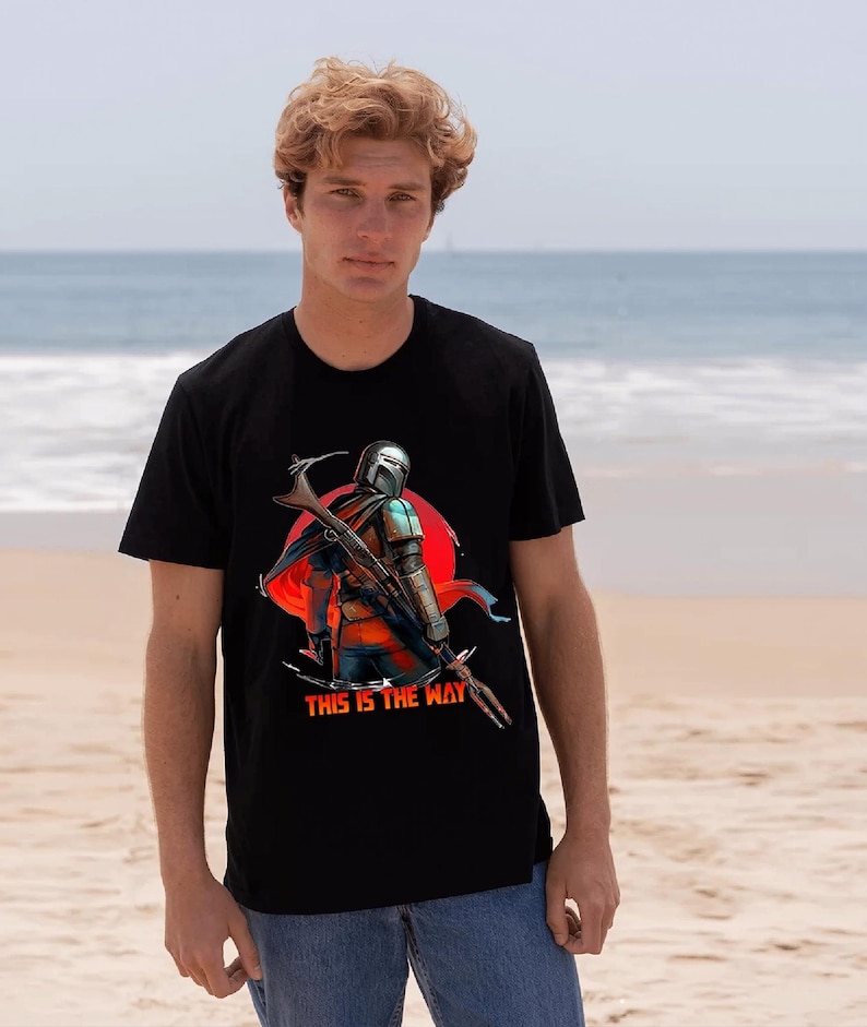 Op de afbeelding: Zwart t-shirt met een afbeelding van een personage met helm en wapen, tegen een rode cirkel. De tekst "THIS IS THE WAY" staat eronder. Het t-shirt wordt door een persoon gedragen.