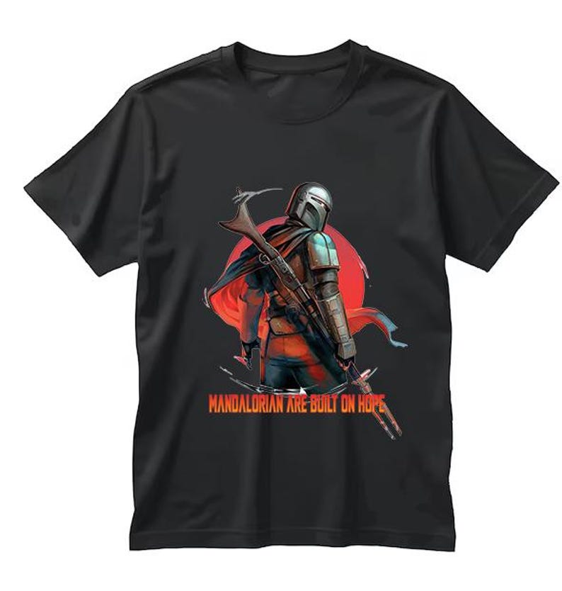 Op de afbeelding: Zwart t-shirt met een gedetailleerde illustratie van een Mandalorian krijger met een blastergeweer, tegen een rode zon. De tekst "Mandalorian are built on hope" staat eronder.