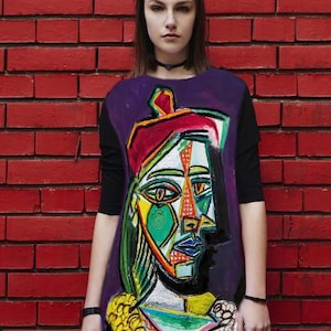 May include: A black short-sleeved tunic featuring a large, colorful, abstract cubist-style portrait print on the front. The graphic includes a face with geometric shapes in shades of green, yellow, red, and blue, set against a deep purple background.