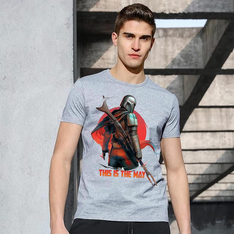 Op de afbeelding: Grijs t-shirt met een afbeelding van een Mandalorian-personage tegen een rode zon. De rode tekst "THIS IS THE WAY" is eronder gedrukt. Het shirt heeft korte mouwen.