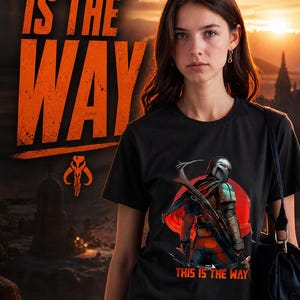 Op de afbeelding: Zwart t-shirt met de tekst "THIS IS THE WAY" in oranje. Het shirt heeft een afbeelding van een personage met een wapen en dezelfde zin. De achtergrond toont een zonsondergang en een ruimteschip.