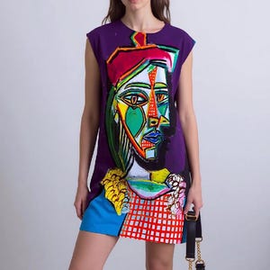 May include: A sleeveless purple mini dress featuring a large, colorful, abstract cubist-style portrait print on the front. The design incorporates vibrant shades of green, red, orange, and blue with a checkered pattern at the hem and a black handbag.