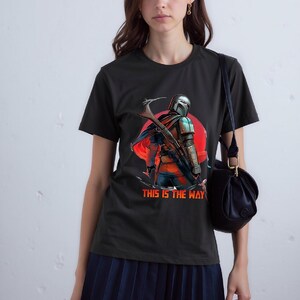 Op de afbeelding: Zwart T-shirt met een afbeelding van een Mandalorian-personage, rode zon achtergrond en de tekst "THIS IS THE WAY". Het ontwerp toont een helm, harnas en een wapen. Het T-shirt is gemaakt van zacht materiaal.