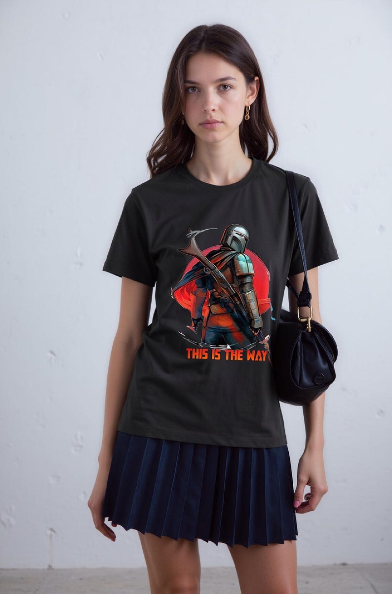 Op de afbeelding: Zwart T-shirt met een afbeelding van een Mandalorian-personage, rode zon achtergrond en de tekst "THIS IS THE WAY". Het ontwerp toont een helm, harnas en een wapen. Het T-shirt is gemaakt van zacht materiaal.