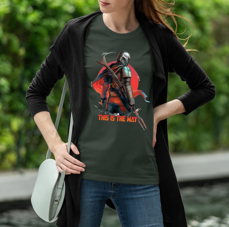Op de afbeelding: Donkergroen T-shirt met een afbeelding van een personage met helm en wapen, tegen een rode cirkel. De tekst "THIS IS THE WAY" staat eronder gedrukt. Het T-shirt wordt door een persoon gedragen.