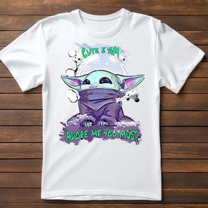 May include: White t-shirt featuring a cartoon illustration of a character with large ears, in shades of green, purple, and blue. The text "Cute I Am" and "Adore Me You Must" are printed above and below the character.