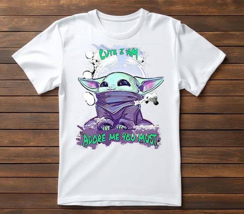 May include: White t-shirt featuring a cartoon illustration of a character with large ears, in shades of green, purple, and blue. The text "Cute I Am" and "Adore Me You Must" are printed above and below the character.