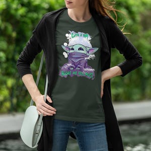 May include: Dark green t-shirt featuring a cartoon character with large ears, a purple cloak, and the text "CUTE I AM" and "ADORE ME YOU MUST". The character is set against a white and green background.