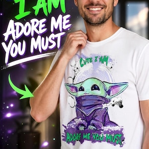 May include: White t-shirt featuring a cartoon character with the text "Cute I Am, Adore Me You Must" in green and purple. The graphic depicts a green and purple alien-like creature. The shirt is worn by a person.
